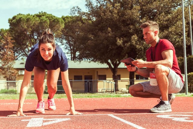 An athletics coach, kneeling with a tablet in hand, watches attentively as his participant assumes a ready position on the starting line, looking ahead with focus.