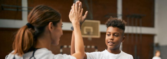 A happy young boy gives his teacher, who has her back to the camera, a high five in the sports hall during a PE session, with other children participating in the session blurred out in the background. 