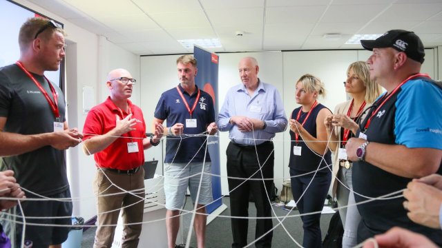 A UK Coaching tutor stands before a group of attentive workshop participants, each holding onto a piece of string that links them together, illustrating the interconnectedness of a game concept