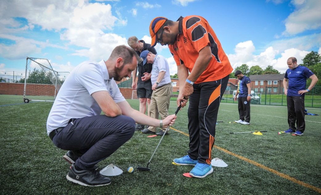 Participants are in a blind golf session. A man in an orange shirt attempts to hit a golf ball while his coach advises on his club position.