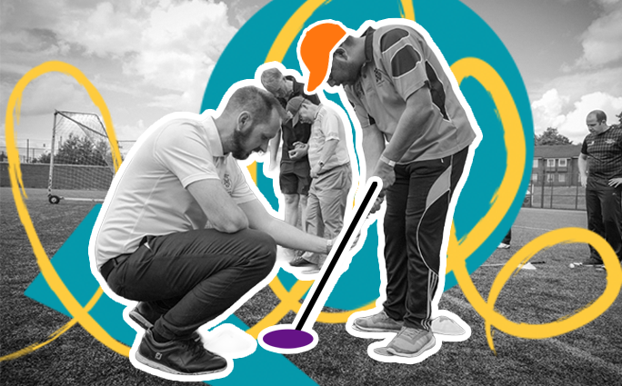 Participants are in a blind golf session. A man in an orange shirt attempts to hit a golf ball while his coach advises on his club position.