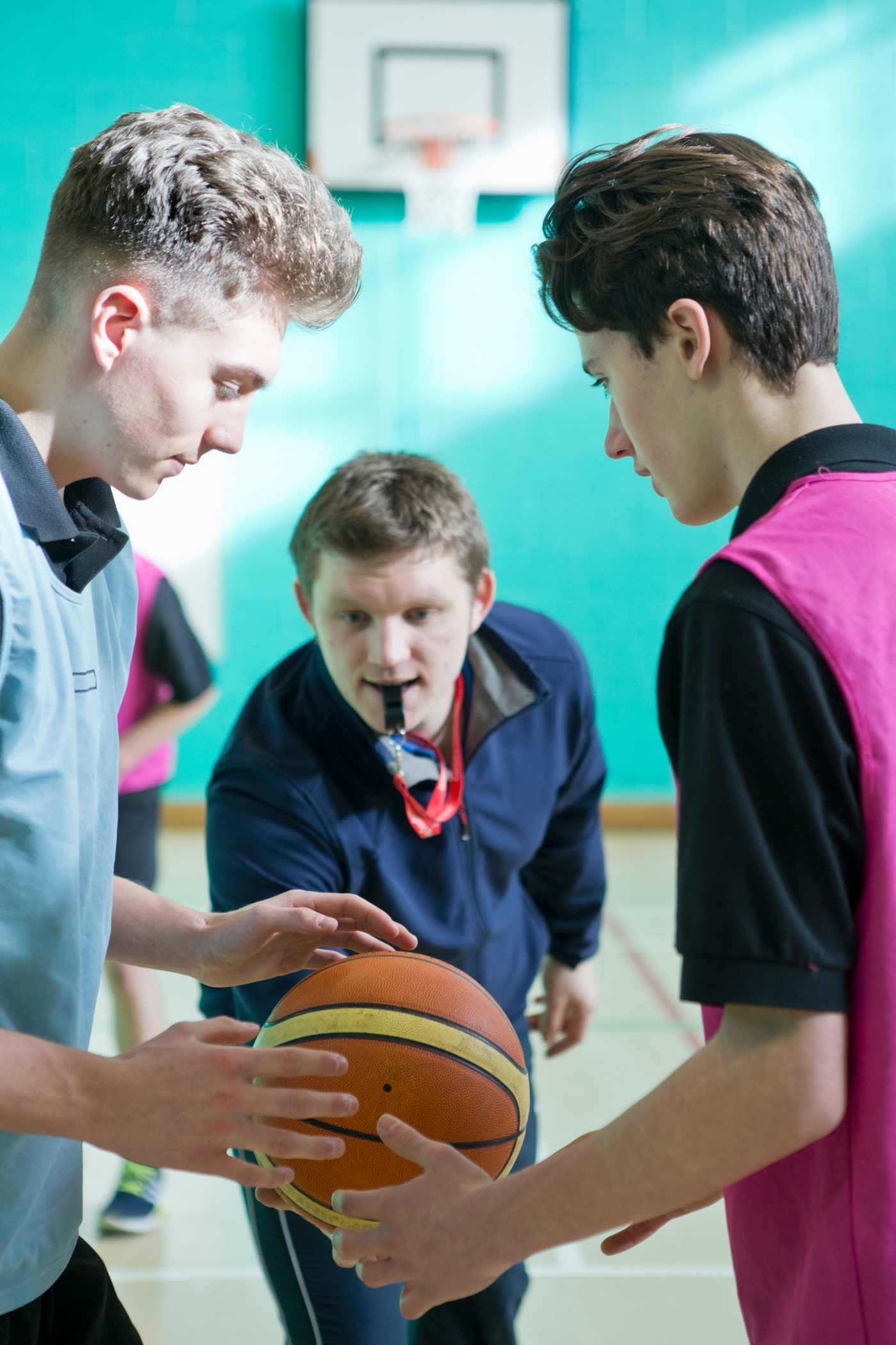 A teacher with a whistle stands between two basketball players, about to throw the ball up in the air for the tip-off.