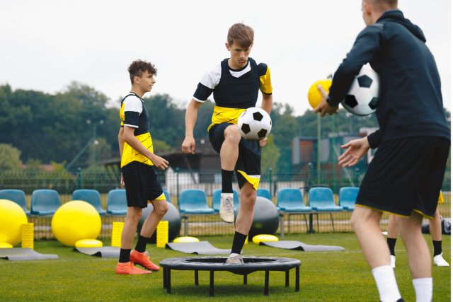 A teenager on a football trampoline skillfully knees the ball back to his coach, who throws footballs at him for agility and control practice, while another teenager watches in the background 