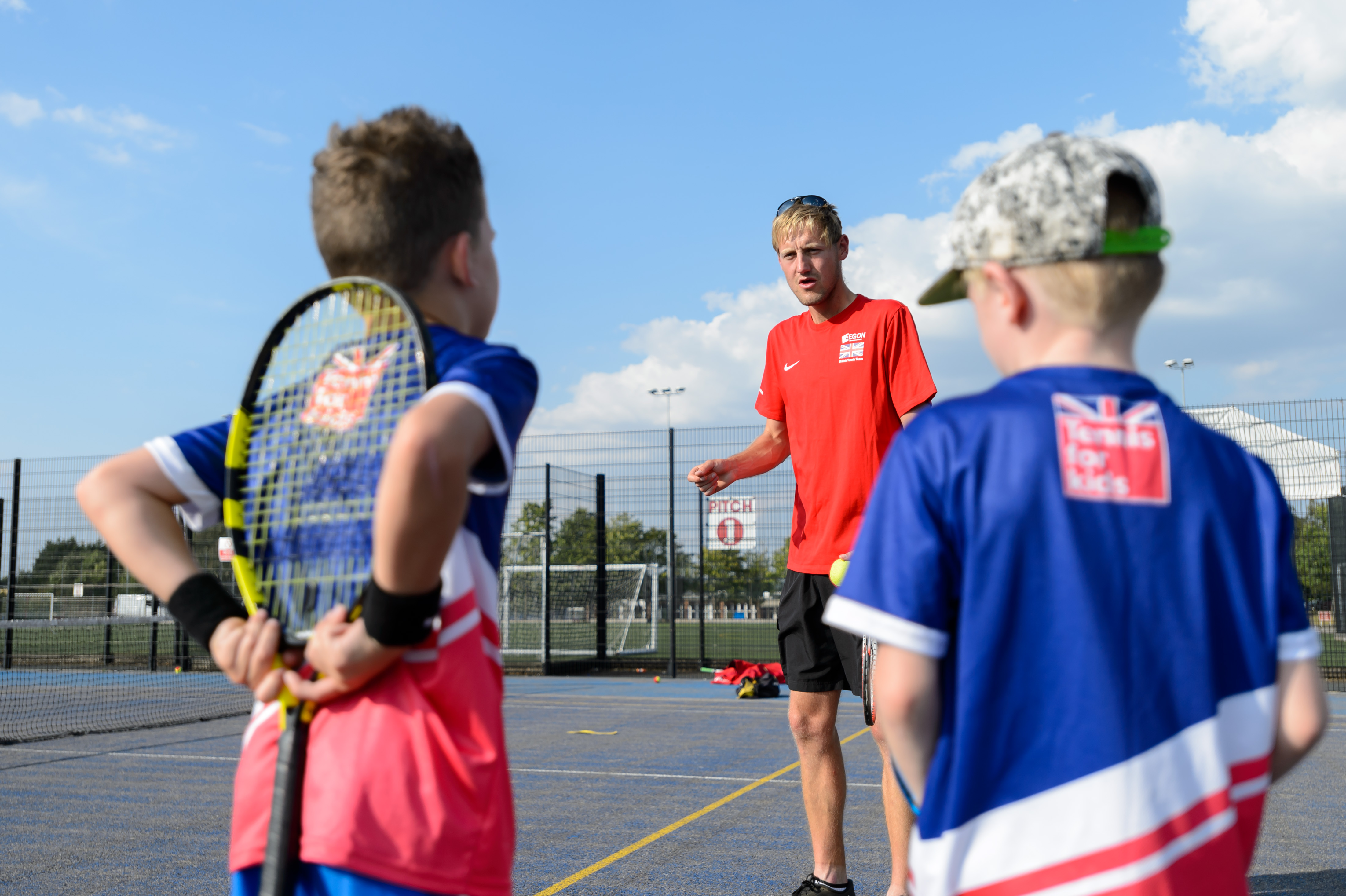 A tennis coach in red talks to two young male participants. One is holding a racket behind their back.