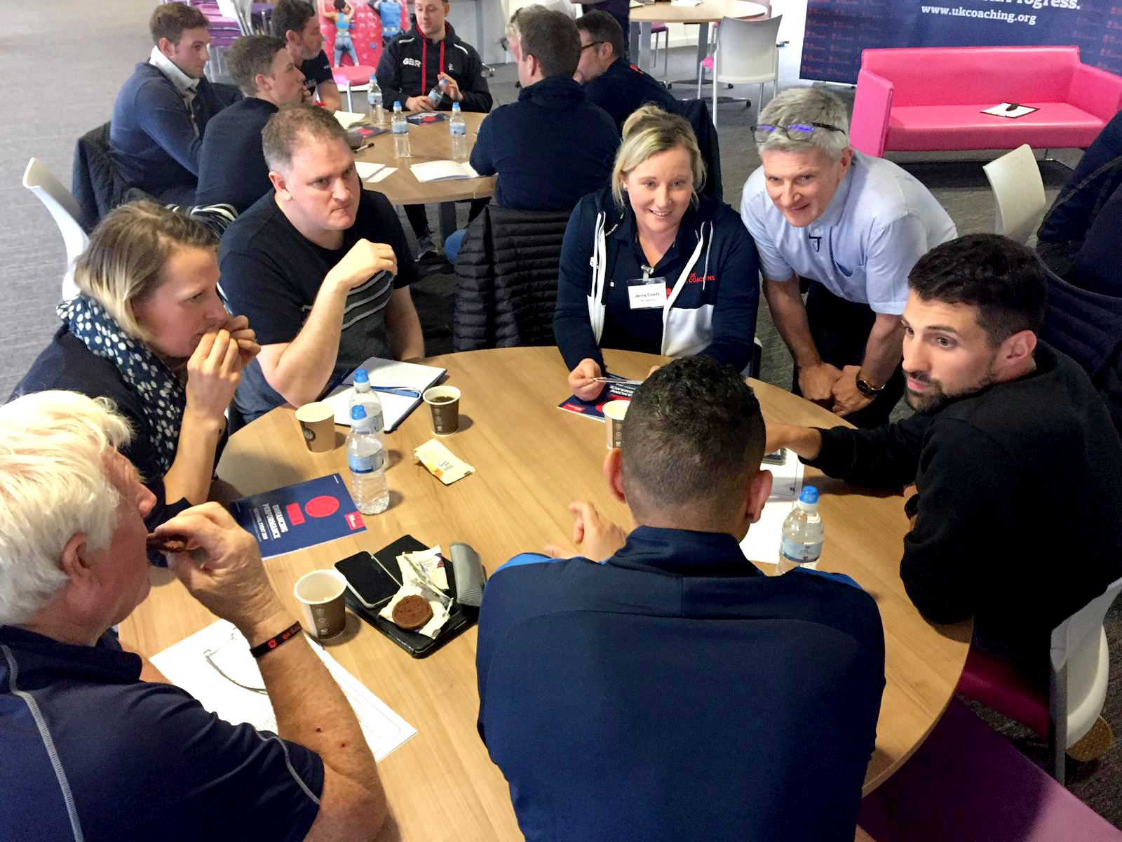 A group of seven coaches sitting around a table in discussion at a UK Coaching branded event.