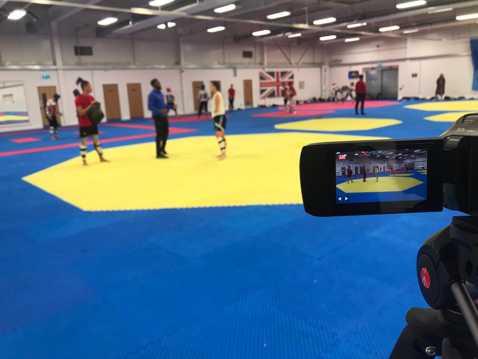 A camcorder is in focus while participants in a martial arts session stand in the background out of focus.