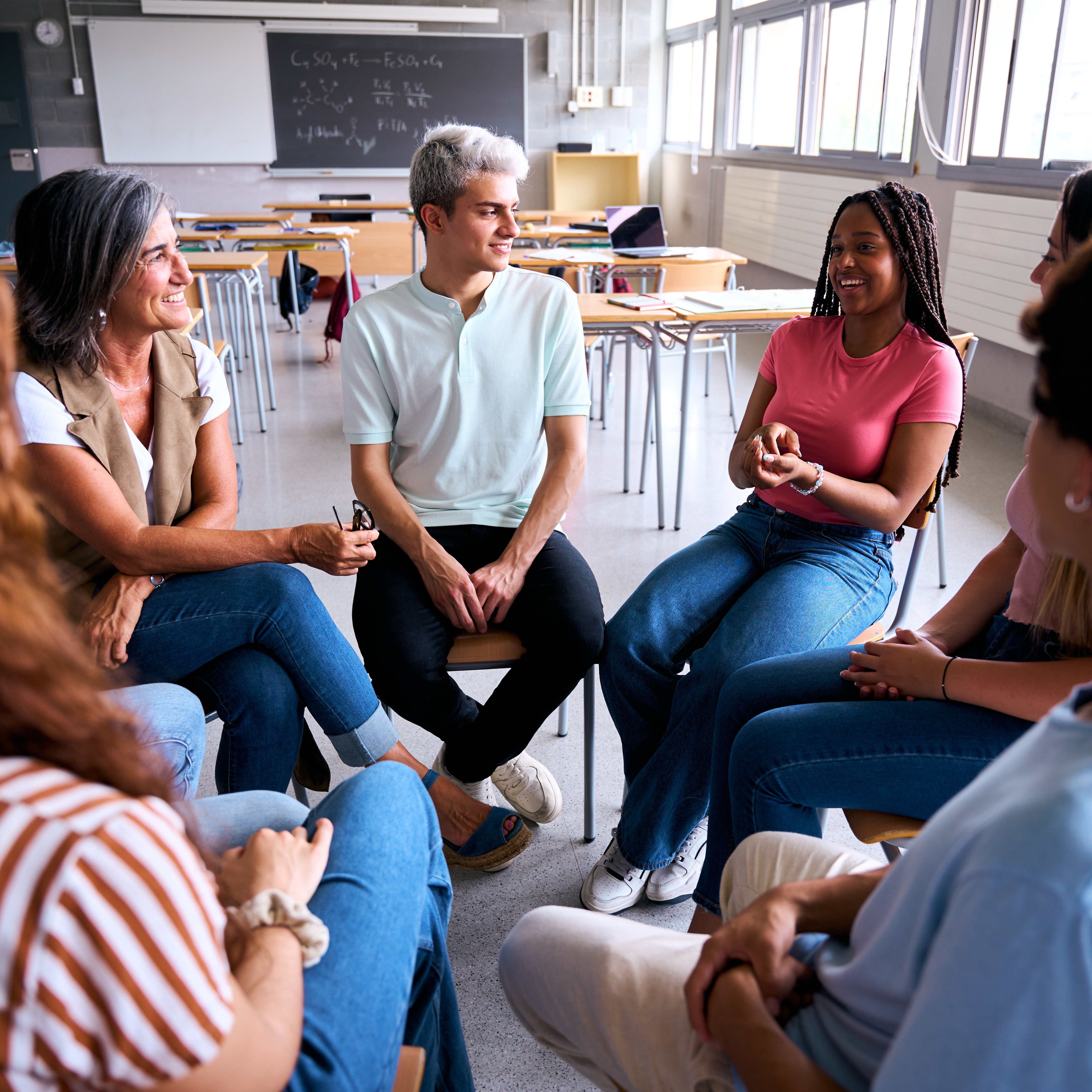 A group of people sit in a circle, listening to a woman in a pink t-shirt talking