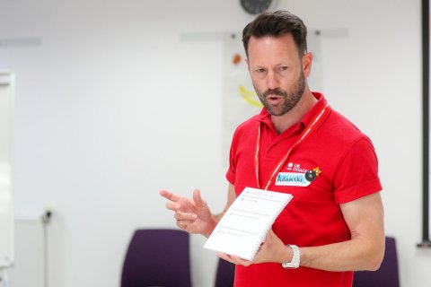 A UK Coaching tutor, wearing a red shirt with a "UK Coaching" lanyard, presenting and gesturing with a notepad in hand.