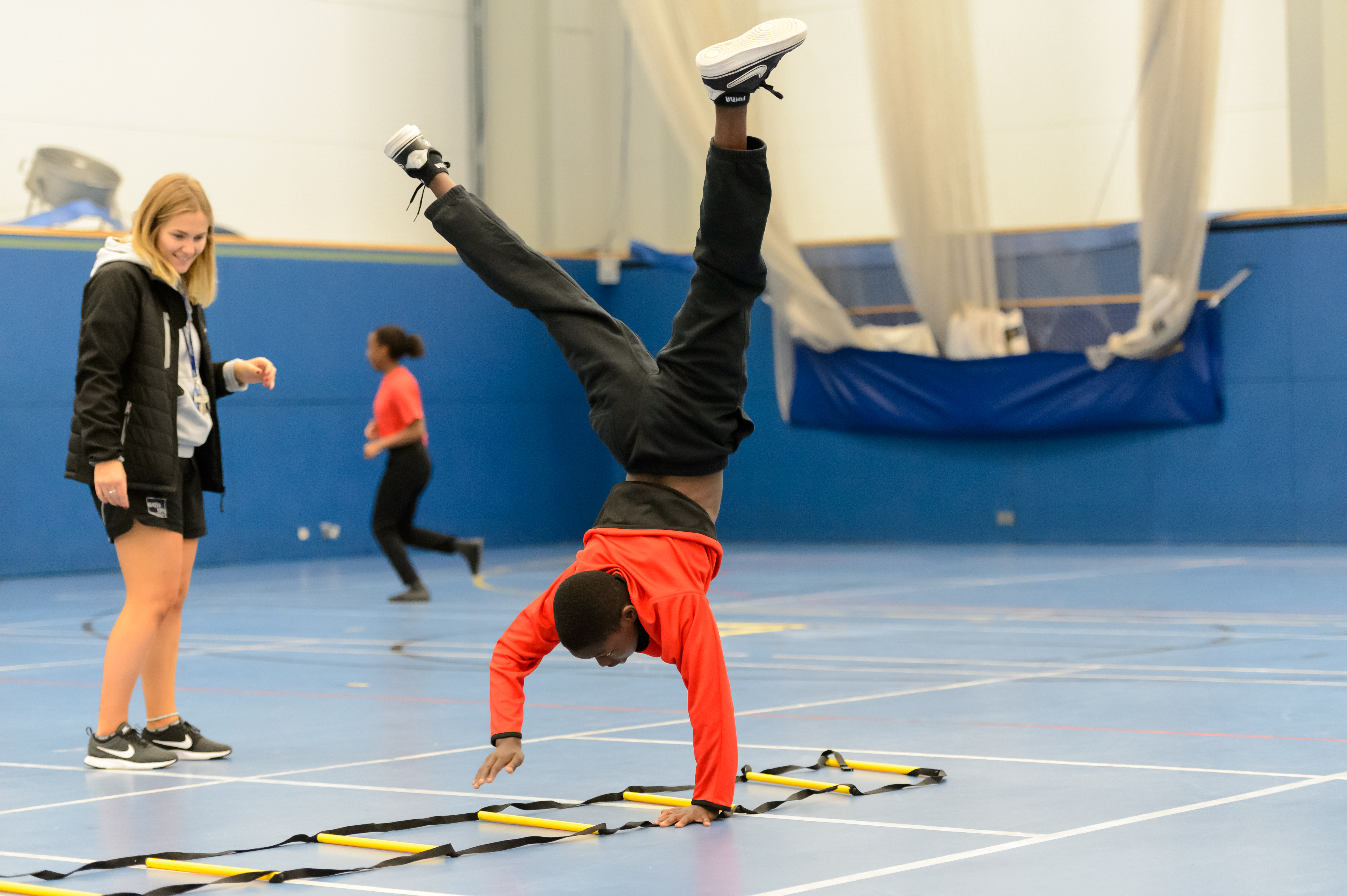 A participant performs a handstand walk while a coach looks on