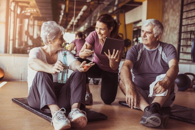 A smiling personal trainer leans over to show something on a tablet to a senior couple she is training. The senior lady smiles back while taking a sip of water, as the man attentively looks on